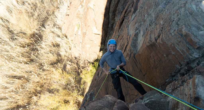 a gap year student who is rock climbing above a desert landscape looks up and smiles